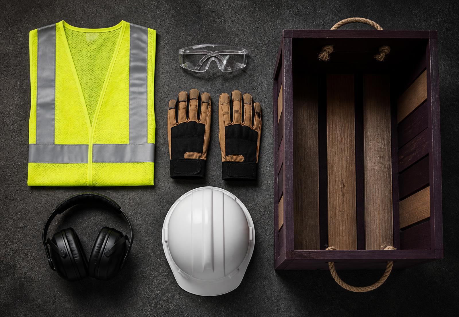 Flat lay of construction PPE — hi-vis vest, safety glasses, work gloves, hearing protection and hard hat — beside a durable wooden work crate